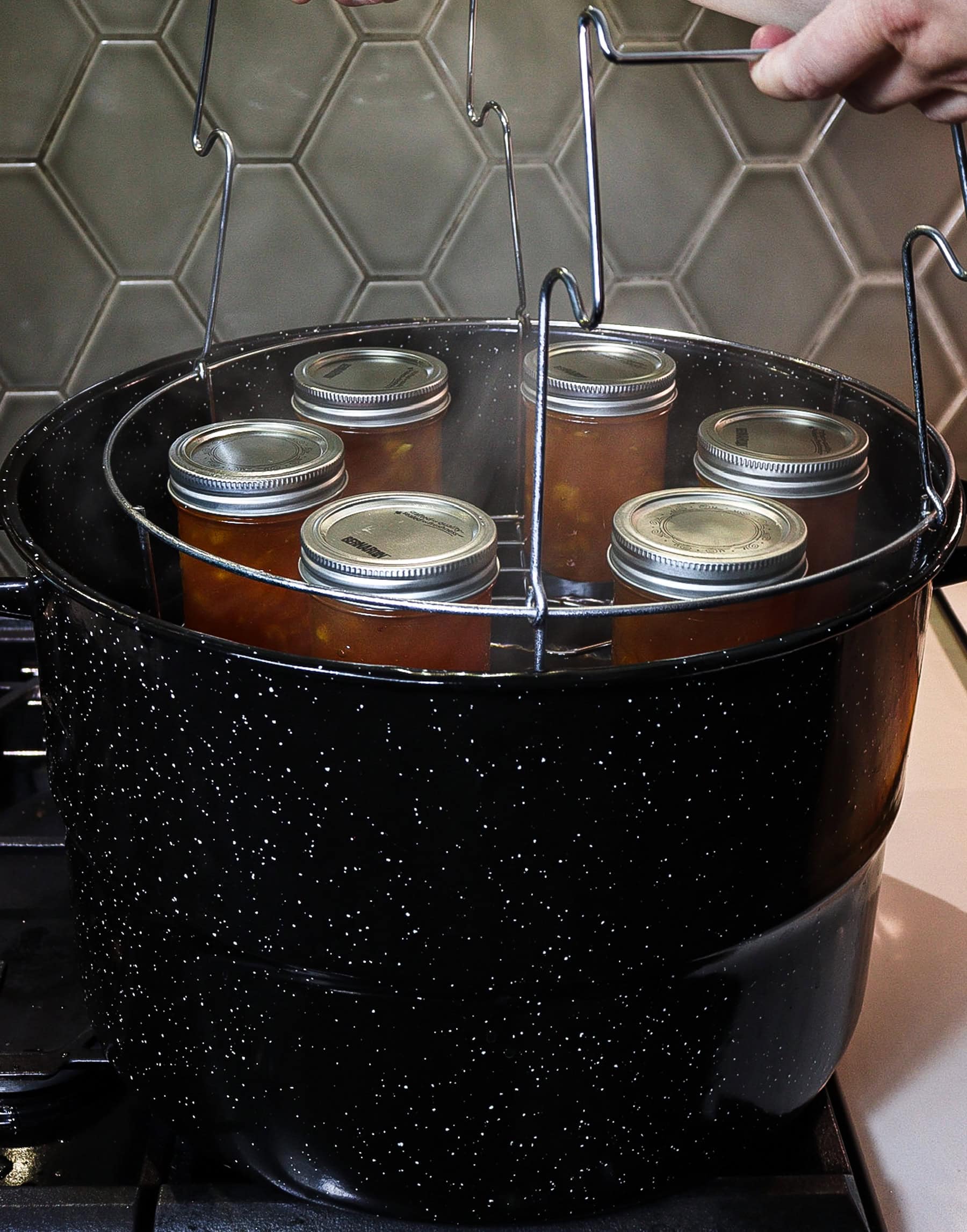 jars of homemade peach jam on a canner rack being lowered into a stovetop pot for a water bath